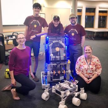 Three highschoolers posing with two library staff members and a model Mars rover